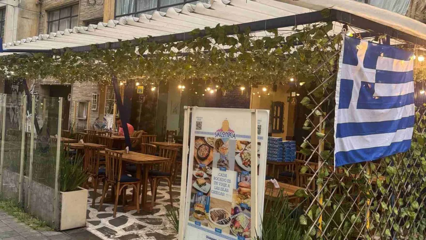 Comedor al aire libre con mesas de madera, letrero publicitario al centro y una bandera griega colgada a la derecha.   