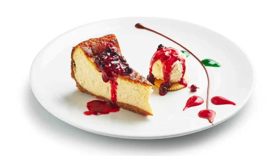 Plato blanco con una tajada de cheesecake bañada en salsa de frutos rojos y una bola de helado decorada con salsa.   