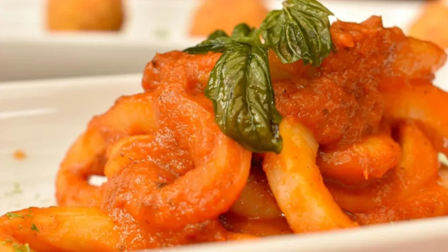 Primer plano de pasta roja con abundante salsa napolitana y hojas verdes de albahaca en plato blanco de cerámica.   