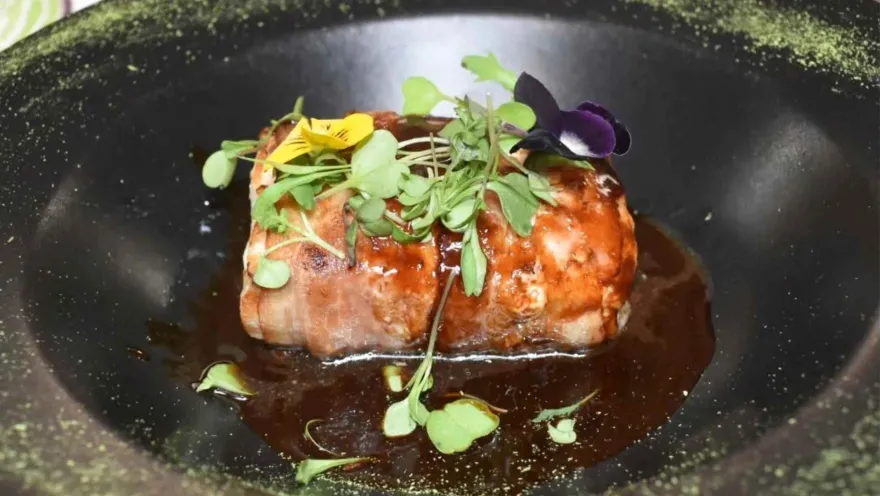 Presentación gourmet de carne en salsa en plato hondo oscuro con germinados verdes y una pequeña flor morada encima.   