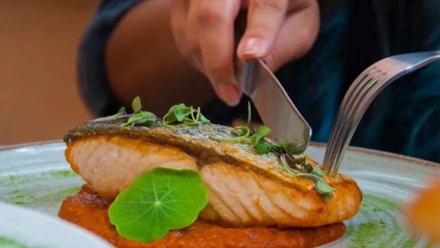 Corte de salmón con piel crujiente servido en plato llano con crema de pimentón, decorado con una hoja verde redonda.   