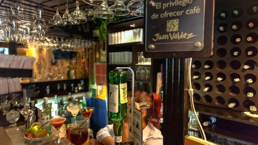 Vista de la barra de atención con cócteles de colores, estantes con botellas y un letrero institucional de café.   