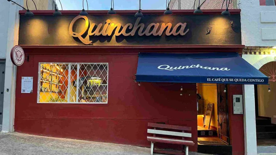 Vista frontal de un café comercial con pared roja, aviso dorado de Quinchana, banca de madera y toldo azul en la calle.