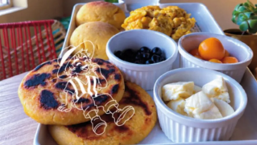 Plato de desayuno tradicional con arepas doradas, recipientes con arándanos, uchuvas y queso junto a panes artesanales.   