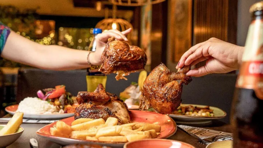 Primer plano de personas tomando piezas de pollo a la brasa dorado sobre un plato con papas fritas y acompañamientos.   