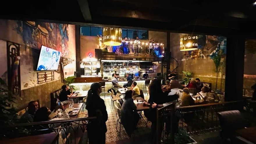 Vista de un bar restaurante iluminado con mesas llenas de personas, lámparas colgantes y tarima superior con músicos.   