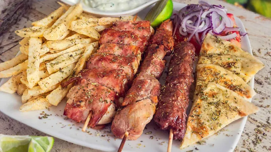 Bandeja blanca servida con chuzos de carne a la parrilla, papas fritas sazonadas y cebolla junto a pan de pita.   