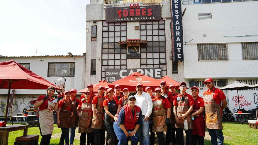 Grupo de colaboradores con delantales marrones y gorras rojas posan sonrientes frente al local Los Torres.