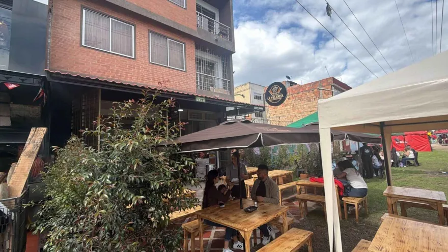 Zona de terraza con mesas de madera, sombrillas oscuras y clientes reunidos frente al letrero de La Velenita.   
