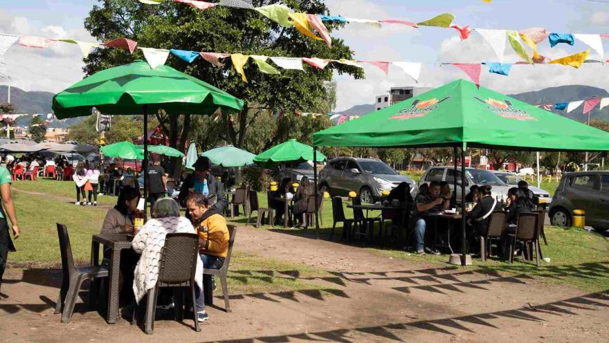 Comensales disfrutando en mesas bajo carpas y sombrillas verdes en una amplia zona verde decorada.