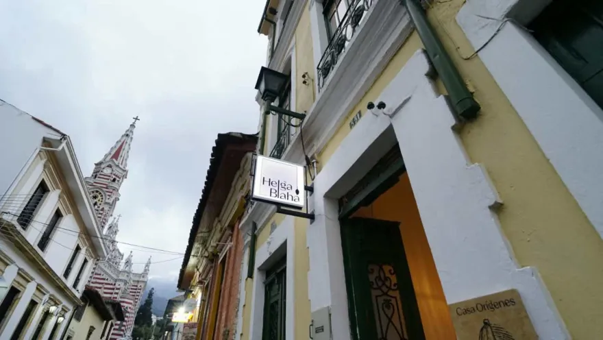 Fachada colonial de paredes amarillas con aviso de Helga Blaha, entrada de madera y la iglesia del Carmen al fondo