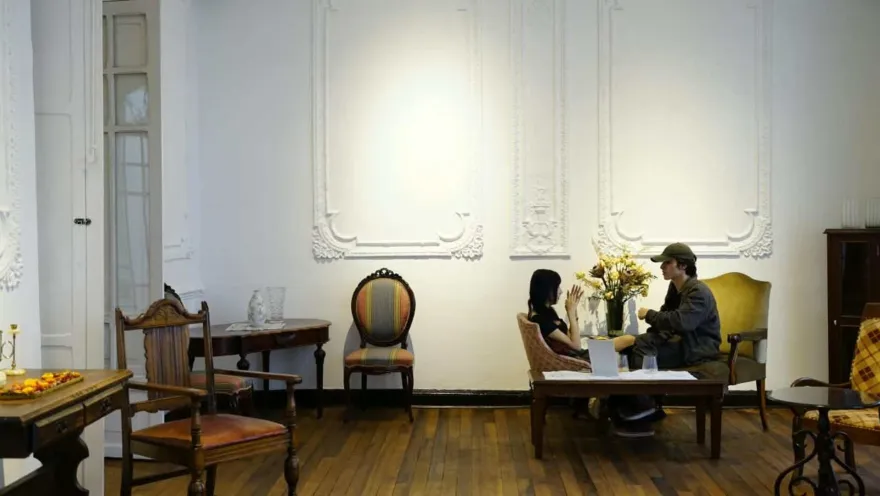 Pareja conversando en una mesa rodeada de sillones antiguos, paredes blancas con molduras y piso de madera rústica.  