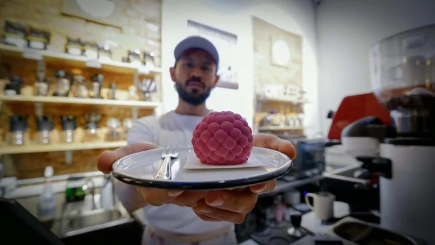 Barista sosteniendo plato blanco con un postre esférico color rosado con textura de frambuesa en la barra del café.  