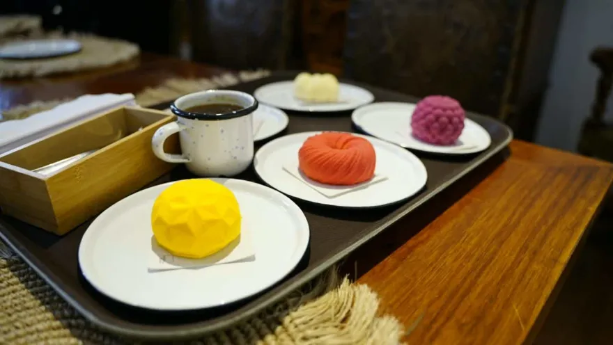 Bandeja servida con tazas de café y platillos con postres geométricos de colores amarillo, rojo, blanco y morado. 