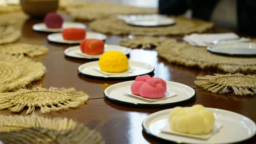 Hilera de platillos con postres artesanales de colores vibrantes sobre mesa de madera decorada con individuales de fibra.