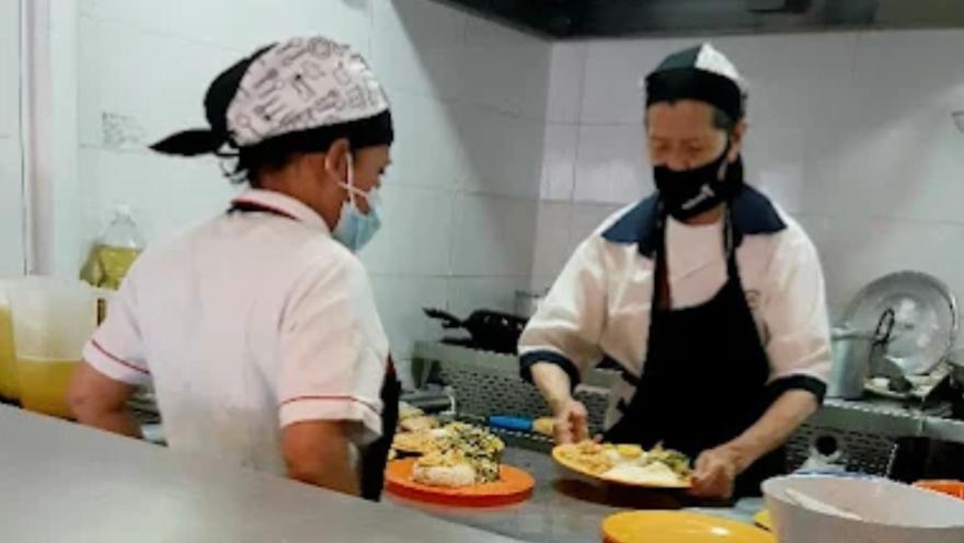  Dos personas con cofia y tapabocas trabajan en la cocina sirviendo platos de comida sobre la barra de atención.