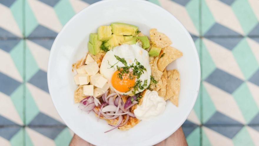 Plato servido en un tazón blanco con nachos, aguacate, huevo pochado, queso en cubos, cebolla morada y crema.