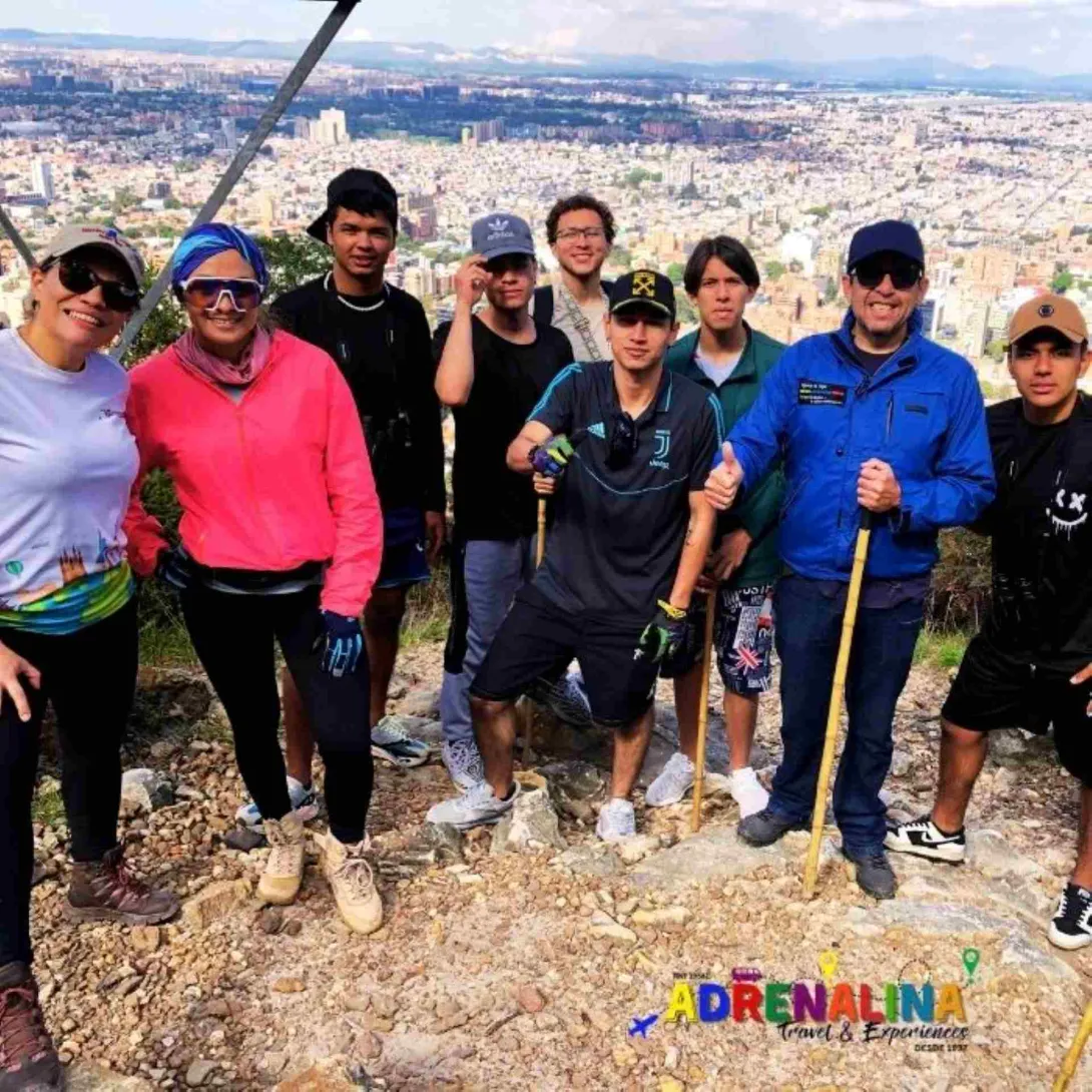  Grupo de personas jóvenes junto con un guía, delante de un mirador donde se observa la ciudad de Bogotá. Todos con ropa deportiva, algunos hombres llevan gorra.