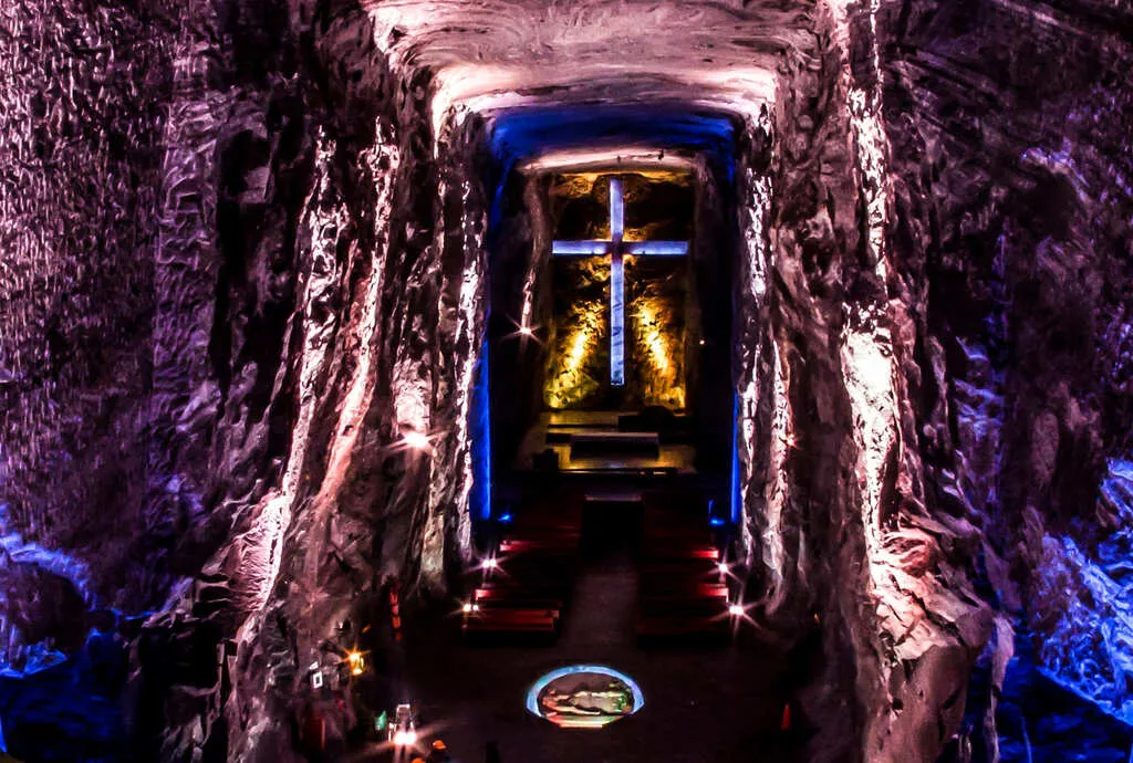 Salt Cathedral of Zipaquirá, inside the cavern illuminated with pink and blue lights, featuring a blue cross with yellow lights in the background.