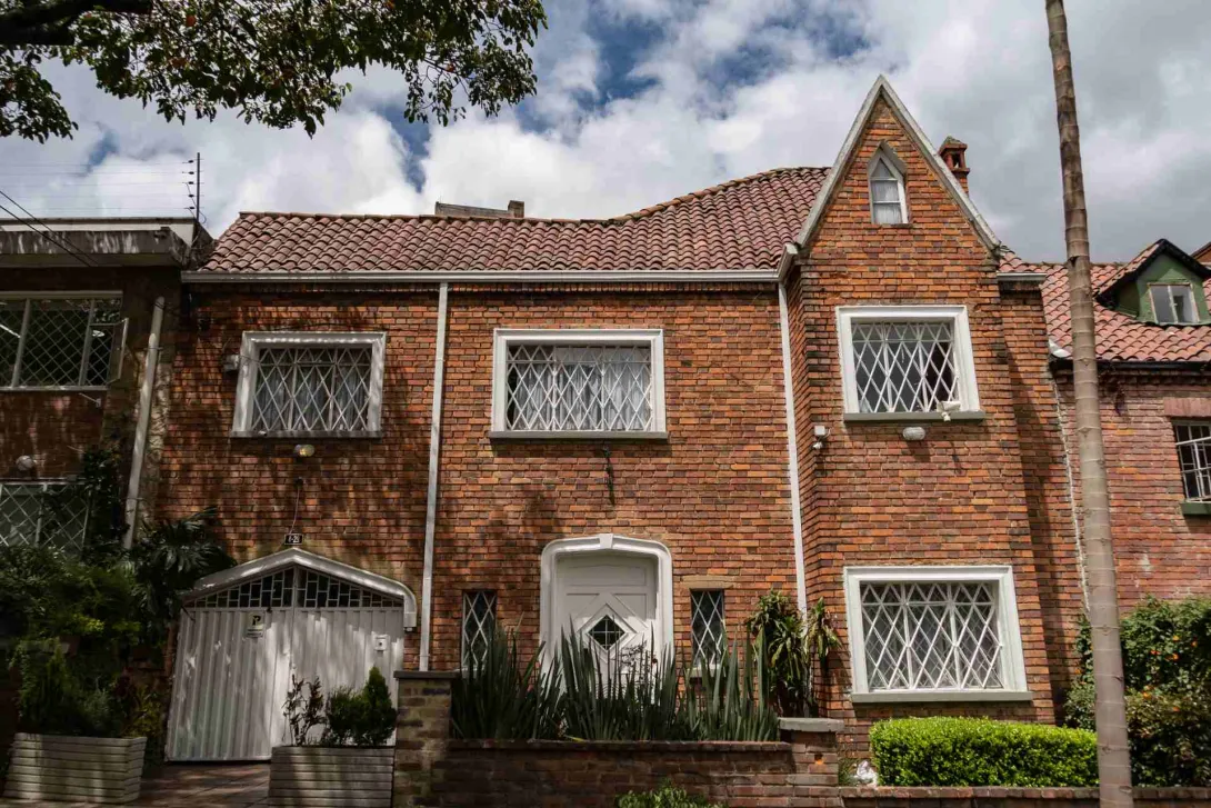 Chapinero primer plano fachada casa estilo inglés en ladrillo, techos en teja de barro y ventanas con marcos y rejas metálicas. La casa cuenta co antenjardín con plantas, asío como la puerta al garaje.