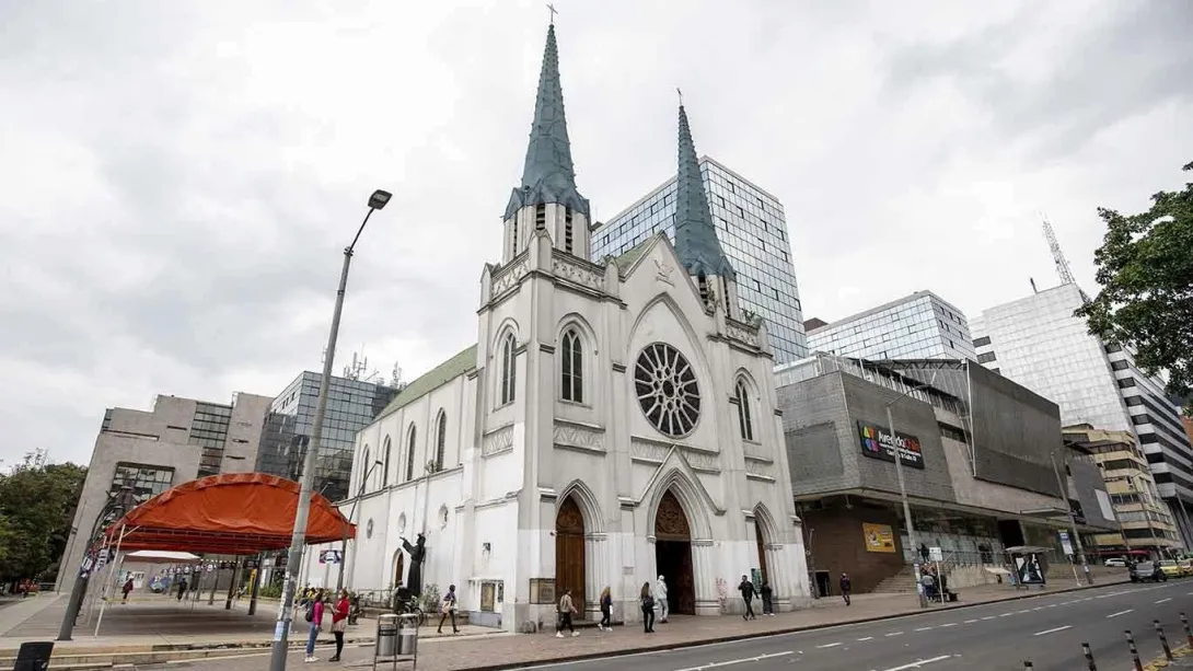 La Porciúncula Church, Gothic-style white facade on Calle 72, with modern buildings and an event tent in front. Several people are walking around. The two gray spires on the roof stand out against the white sky.