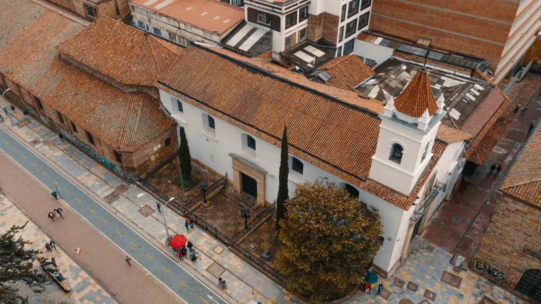  Iglesia de La Veracruz colonial su techo con tejas de barro y una torre blanca con el campanario recubierto con teja y una cruz metálica que lo corona. Se obsera la carrera séptima su cicloruta con algunas personas caminando y un par de puestos de vendedores ambulantes.