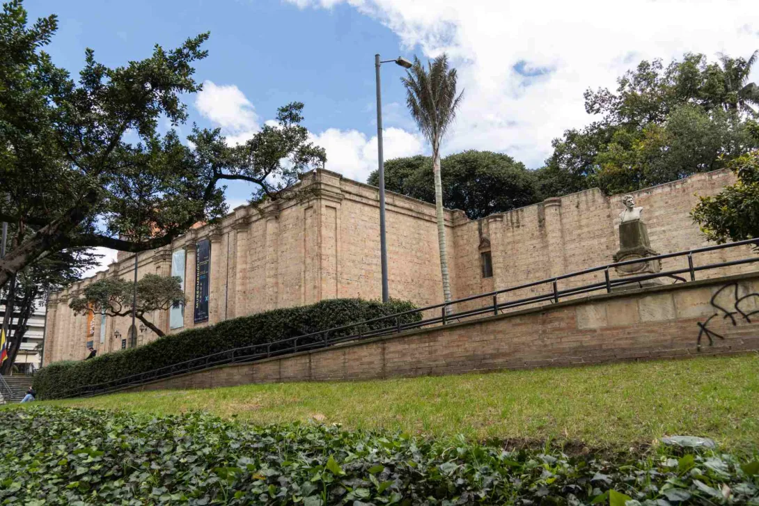 National Museum of Colombia, wide shot of the façade seen from below in the left corner, surrounded by vegetation, trees, and a tall palm tree. Above the stone building, the sky is blue with clouds.