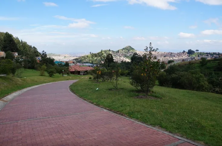 Parque Entrenubes con camino de ladrillo rodeado por árboles pequeños y otra vegetación, al fondo se ven montañas y mucho más lejos la ciudad. El cielo está azul con algunas nubes.  