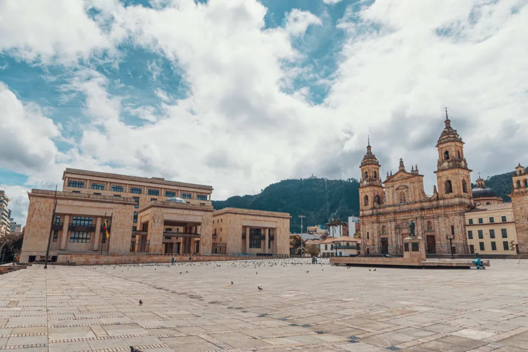 Plaza de Bolívar de día vista de esquina suroccidente al norte con palomas pequeñas a lo lejos, de frente el Palacio de Justicia, de lado la Catedral Primada, al fondo el cerro de Monserrate y el celo azul muy nubado.