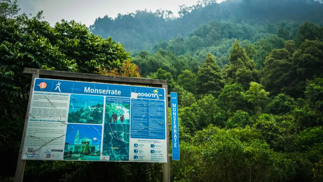 Sendero Cerro Monserrate con aviso en madera con titulación de Monserrate rodeado de árboles frondosos en una montaña. 