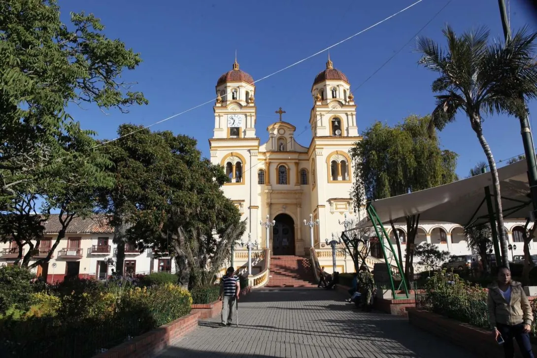  Basílica Menor de San Jacinto fachada de frente vista desde la paza rodeada de árboles y una palma, con un hombre con bastón y una mujer que caminan alejándose de ella. El cielo es muy azul.