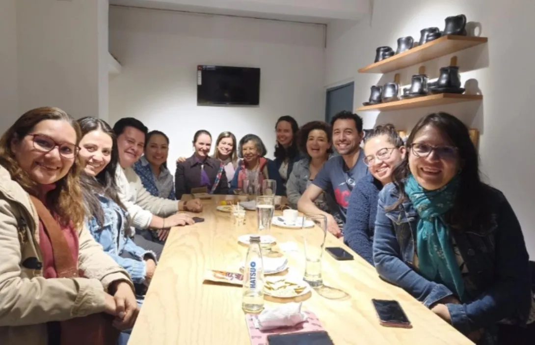 Grupo de personas en un salón, mayoría mujeres jovenes y adultas ríen mirando al frente. Al lado en una repisa en la pared hay unos tazones negros de chocolate.
