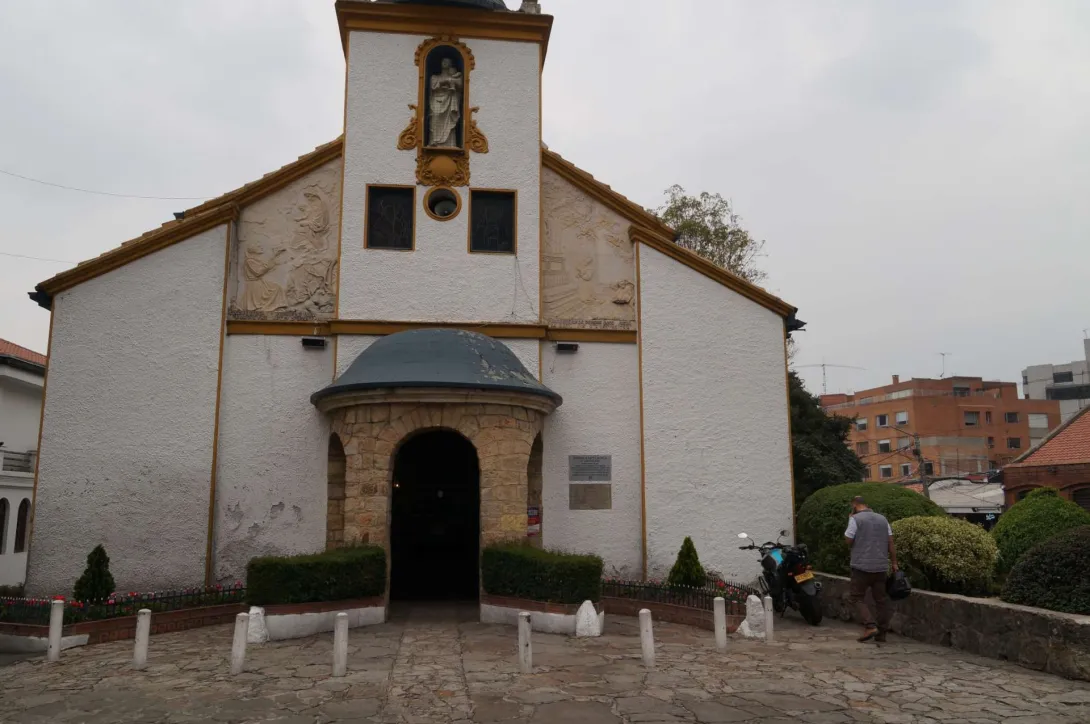 Iglesia Santa Mónica plano general fachada con un hombre adulto y una moto estacionada a la izquierda. Al frente el piso es en piedra y la puerta de la entrada está cerrada. al fondo a la izquierda aparece un edificio moderno.