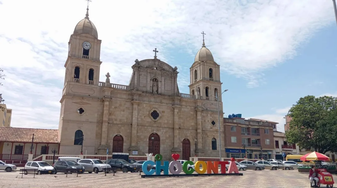 Our Lady of Health Parish - Chocontá, wide shot of the façade during the day, with a blue sky and several cars in front, featuring the large billboard with the name Chocontá.