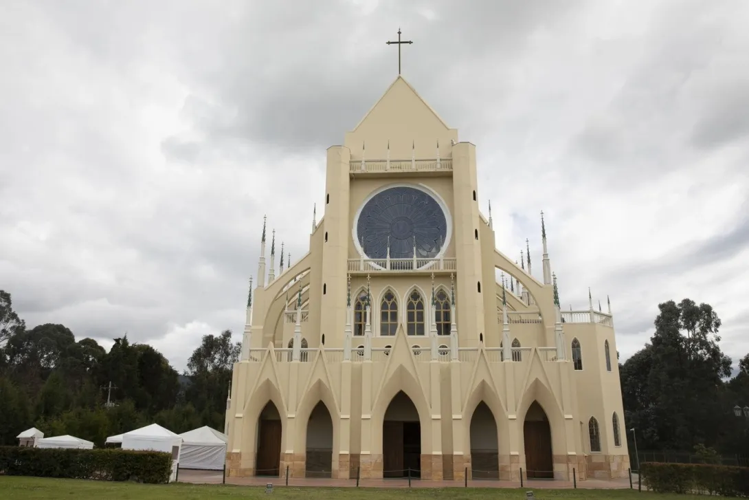 Santuario Nuestra Señora de Fátima - Tocancipá fachada gótica en primer plano coronada por una cruz metálica y con árboles frondosos alrededor y un cielo gris. 