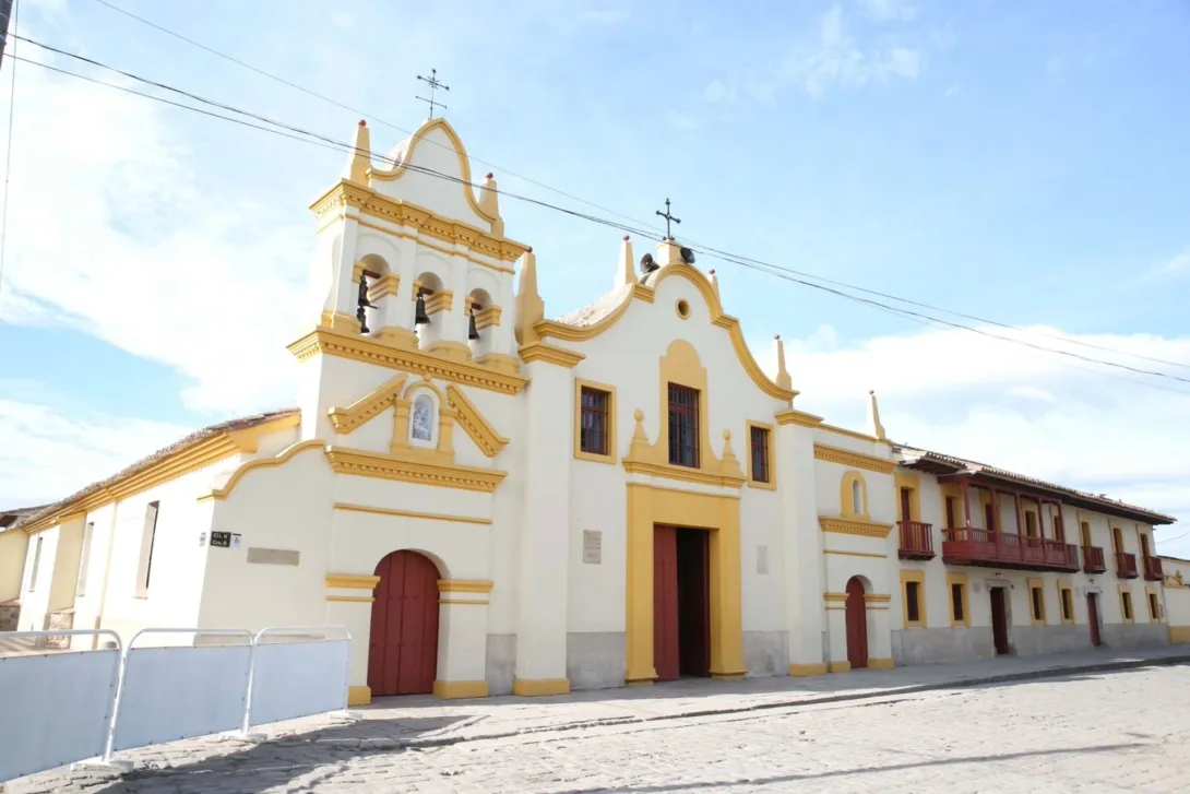 Sanctuary of Our Lady of Health - Bojacá, wide diagonal view of the façade with colonial houses on the left side. The façade is white with yellow stripes, and the bell tower with four bells stands out. The sky is blue with a few clouds.