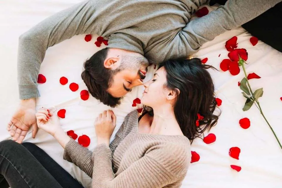 Pareja de hombre y mujer jóvenes recostados sobre una cama decorada con rosas rojas. 