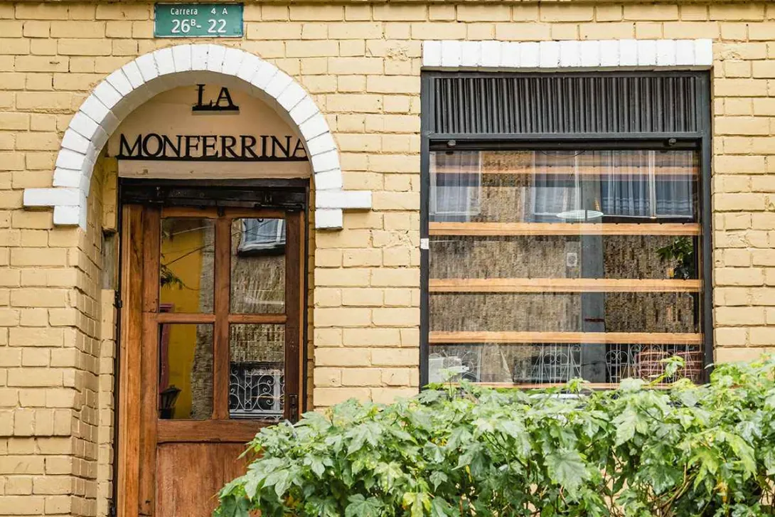 Esta fotografía de cerca muestra la fachada de "LA MONFERRINA", un restaurante en el barrio de La Macarena. La arquitectura tradicional del lugar se destaca por su fachada de ladrillo amarillo y una puerta de madera de estilo rústico enmarcada por un arco blanco. El toque de naturaleza lo da un seto de plantas verdes en la parte inferior, que le da vida y color al lugar.