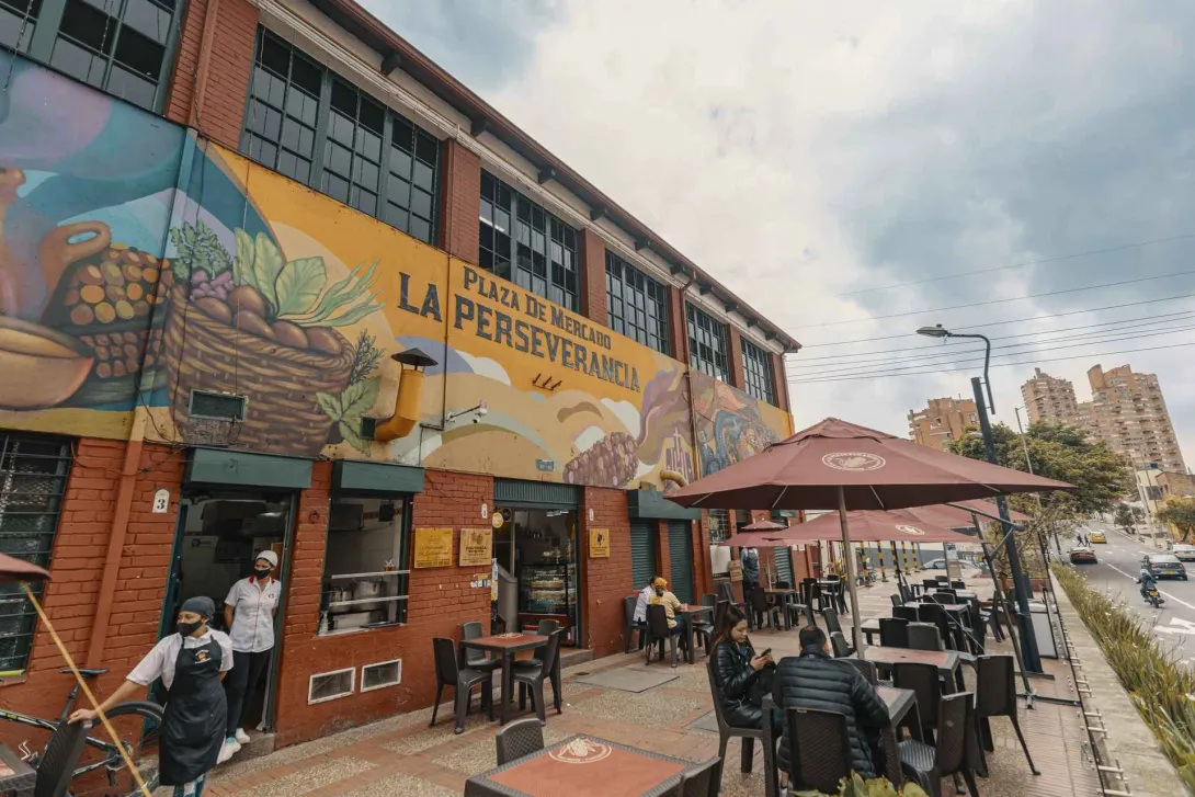 Esta es una vista exterior de la "Plaza de Mercado La Perseverancia", un edificio de ladrillo rojo con grandes ventanas en el piso superior. La fachada está adornada con un vibrante mural que representa escenas de mercado y paisajes. Delante del edificio, hay una zona de mesas y sillas con sombrillas de color marrón, donde varias personas están sentadas. En el lado izquierdo, dos personas con mascarillas, una de ellas con un delantal, están en la entrada. A la derecha, se ve una calle con tráfico y edificio