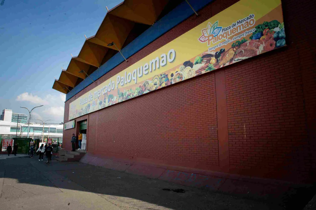  La imagen muestra la fachada exterior de la "Plaza de Mercado Paloquemao" bajo un cielo despejado. El edificio tiene una estructura de ladrillo rojo y una marquesina distintiva con un patrón en zigzag en la parte superior. Un gran letrero amarillo horizontal exhibe el nombre del mercado y un colorido mural con ilustraciones de frutas, verduras, carnes y pescados, junto con el eslogan "Siempre lo mejor, siempre fresco". Varias personas se ven entrando y saliendo de la entrada principal a la izquierda.