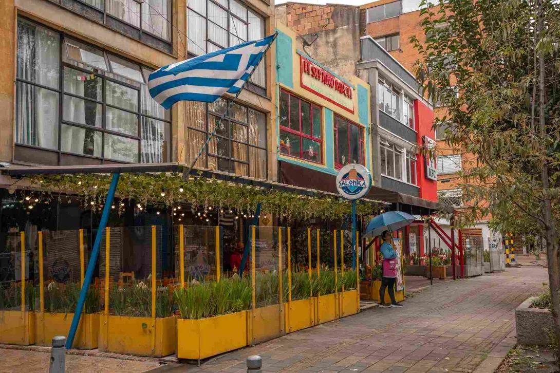 Esta imagen muestra una calle vibrante y llena de color en el Park Way. En primer plano, la fachada de un restaurante de comida griega llamado 'Salonika´ se destaca por sus colores alegres. Un toldo de metal y vidrio con luces colgantes cubre la terraza, creando un ambiente acogedor. La bandera griega ondeando sobre el restaurante y los detalles en amarillo y azul evocan la cultura mediterránea.