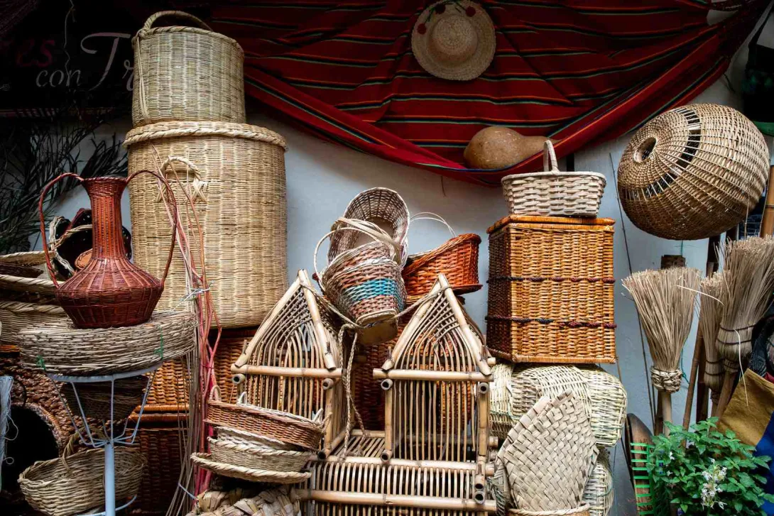 Esta imagen muestra una densa exhibición de productos artesanales hechos de mimbre y materiales naturales. Se observan cestas de diferentes formas y tamaños apiladas, algunas con tapas, y otras con diseños intrincados. También hay objetos decorativos como jaulas de bambú, un jarrón de barro, y manojos de escobas. Un sombrero de paja cuelga sobre una tela roja con rayas en el fondo, creando una atmósfera rústica y tradicional.