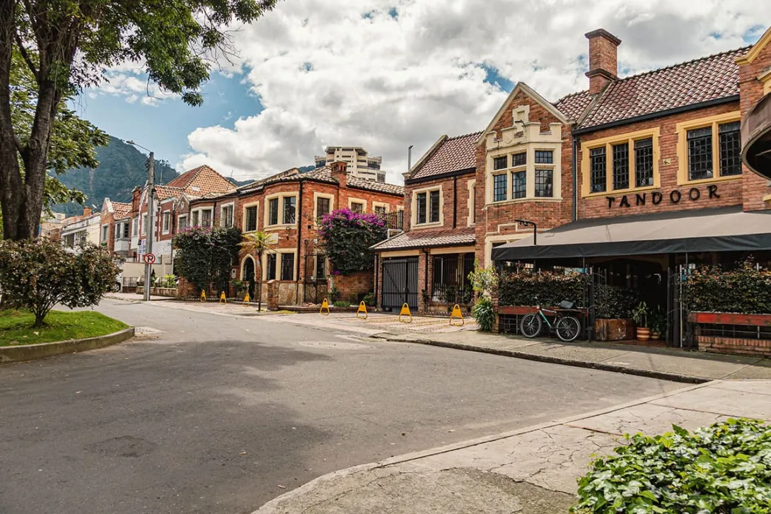 Esta imagen captura la esencia de la Quinta Camacho, un barrio de Bogotá caracterizado por su arquitectura de estilo inglés. Se aprecian varias casas de ladrillo rojo, con techos a dos aguas y ventanas enmarcadas, que se alinean a lo largo de una calle tranquila y arbolada. El ambiente es sereno y residencial, con un cielo parcialmente nublado que realza los colores de las fachadas y la vegetación.