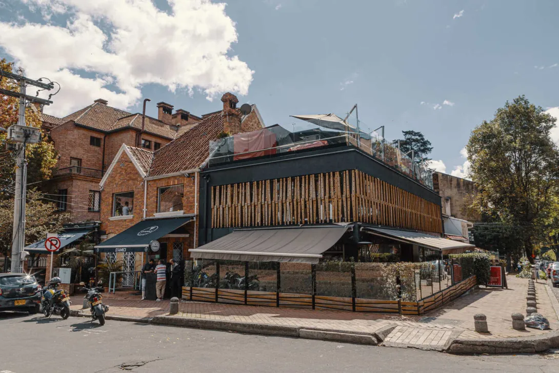 Esta imagen muestra una esquina vibrante de la Zona G en Bogotá. En el centro de la toma, un edificio de dos pisos destaca por su moderna fachada negra con detalles en madera, lo que le da un toque sofisticado y orgánico. El cielo azul con nubes dispersas ilumina el paisaje, creando una atmósfera acogedora.