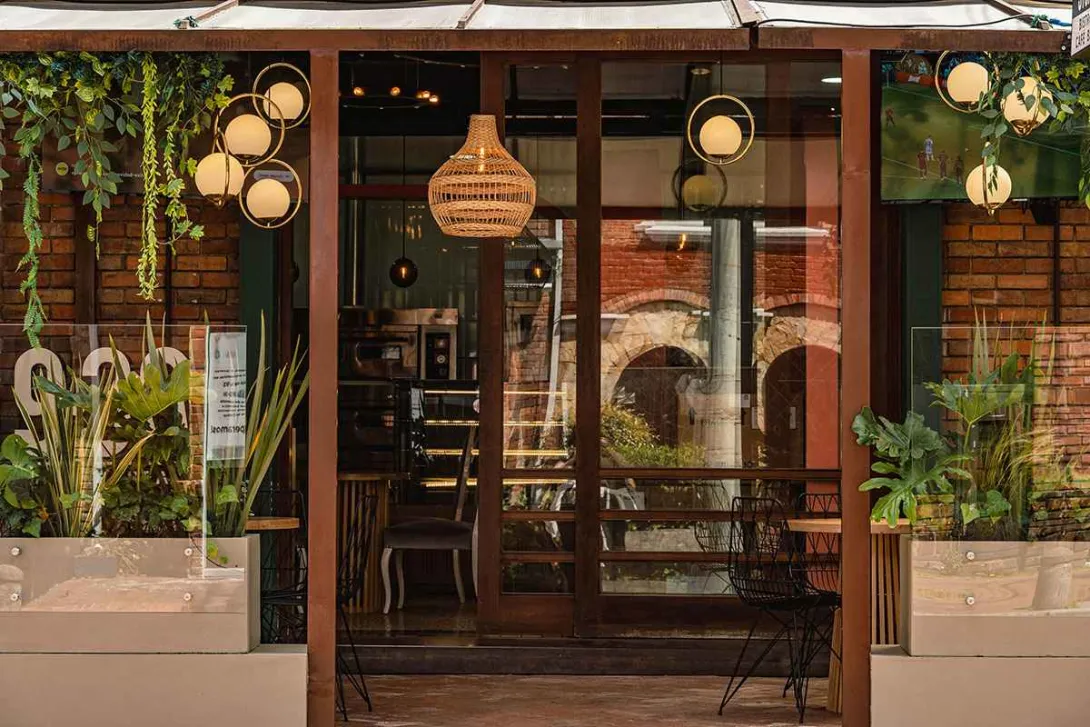 This image shows the facade of a cozy restaurant in Bogotá's Zona G. The entrance, with its large wooden windows, invites you to look inside, where warm lighting and modern decor abound. On either side, potted green plants and hanging lights in the shape of spheres and woven baskets create a sophisticated and natural ambiance. The design integrates rustic elements with contemporary touches, making it an ideal place to enjoy a gastronomic experience in an elegant and relaxed setting.