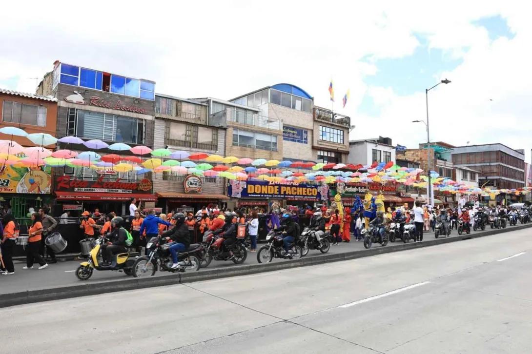 Esta imagen vibrante muestra una calle animada en un sector comercial, posiblemente durante un festival o evento. Una multitud de personas, algunas en motocicleta y otras a pie, se congregan bajo un cielo de paraguas de colores que cuelgan sobre la calle. Los edificios de fachadas variadas, algunos con letreros de negocios locales, crean un fondo dinámico. La presencia de motocicletas y la gente en movimiento sugieren una celebración o un día de actividad comercial intensa, con un ambiente festivo 