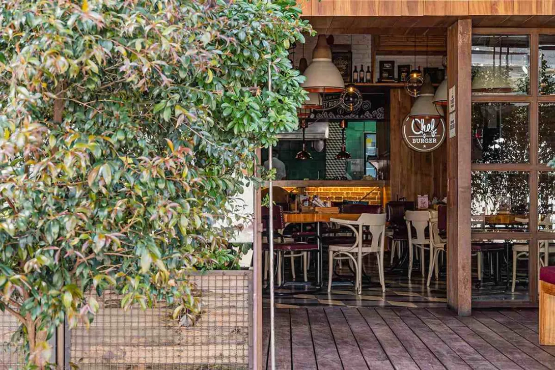 La fotografía muestra la entrada de un restaurante llamado "Chef Burger" en el Parque de la 93. La arquitectura del lugar es moderna y acogedora, con un exterior de madera y grandes ventanales que permiten ver el interior. Las lámparas de estilo industrial cuelgan sobre las mesas, creando un ambiente cálido e íntimo. El diseño del espacio invita a los transeúntes a entrar y disfrutar de una comida en un ambiente relajado y contemporáneo.