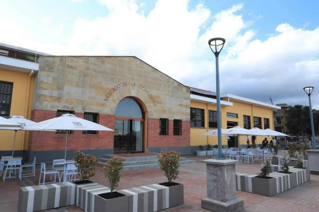 Esta imagen muestra la fachada principal del "Mercado de la Concordia" en Bogotá, con un estilo arquitectónico que combina lo tradicional con lo moderno. La entrada, con su arco de ladrillo y una estructura de piedra en la parte superior, tiene un letrero que lleva el nombre de la plaza. En el exterior, se ha creado una plaza con mesas y sillas blancas bajo sombrillas, que invitan a los visitantes a disfrutar de un espacio abierto y acogedor.