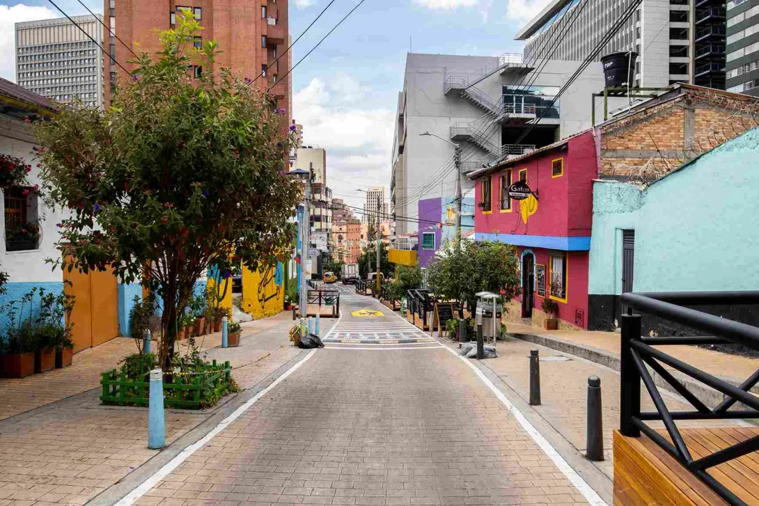 This image offers a wider view of Bogotá’s colorful streets, showcasing how vibrant buildings contrast with the modern architecture in the background. Brick and concrete houses blend seamlessly with facades painted in fuchsia, light blue, and yellow. Lush trees and flower boxes add a touch of nature to the urban landscape. The street, with its brick paving and wooden railings, invites visitors to walk and explore this corner filled with charm and vitality.