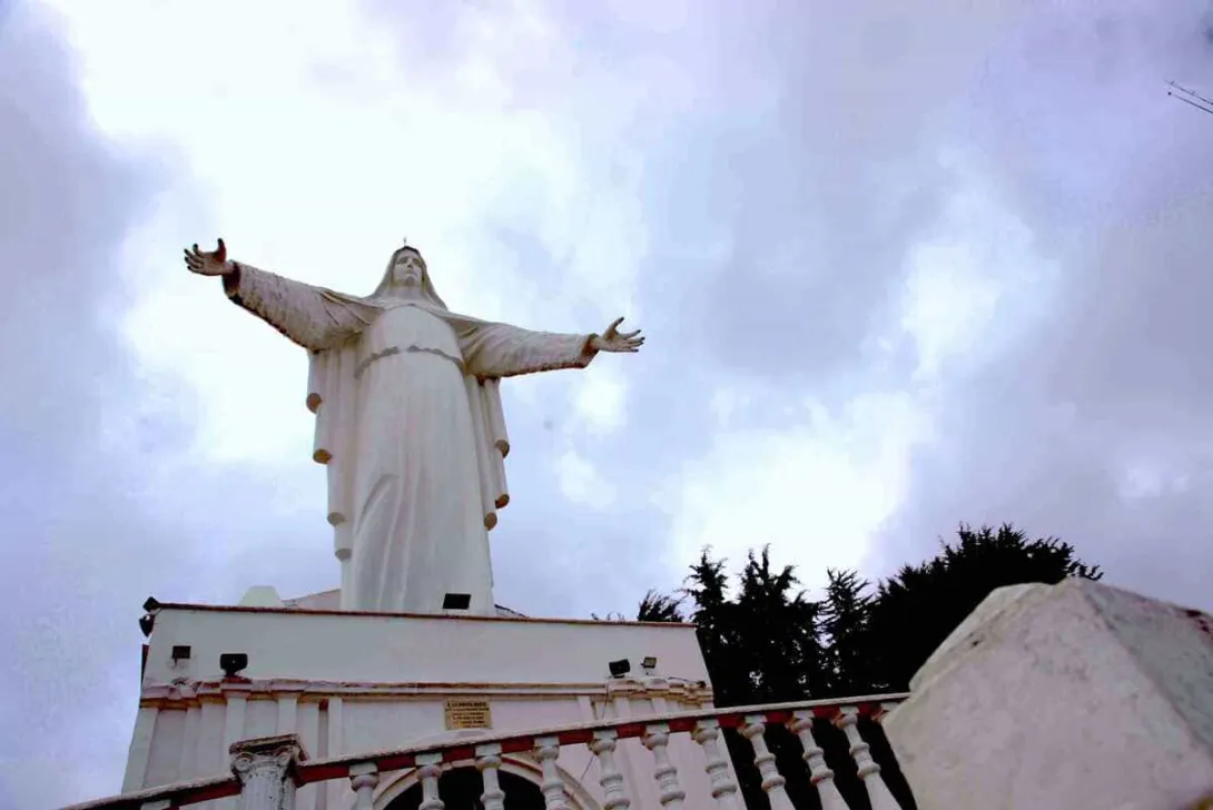 White statue of the Virgin of Guadalupe with open arms, above the church and under a blue sky with dark clouds. It is surrounded by tree tops.
